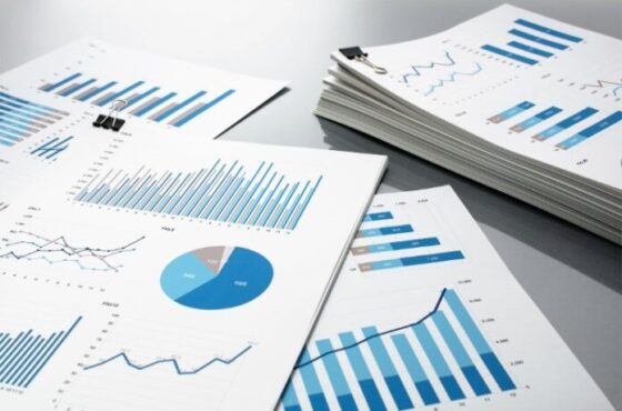 Desk with multiple printed reports showing blue bar charts, line graphs, and a pie chart clipped with binder clips.