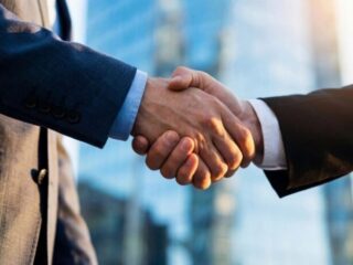 Two businesspeople in suits shake hands in front of a glass office building, signaling an agreement.
