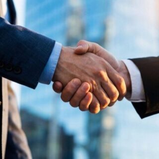 Two businesspeople in suits shake hands in front of a glass office building, signaling an agreement.