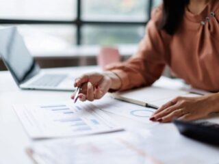 Person in a peach blouse analyzes financial charts on a desk, pointing with a pen beside a laptop.