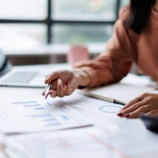 Person in a peach blouse analyzes financial charts on a desk, pointing with a pen beside a laptop.