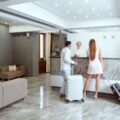 Two guests with rolling suitcases check in at a modern hotel reception behind a marble counter.