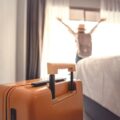 Orange suitcase in the foreground of a hotel room with a person raising their arms by the window in the background, bed on the right side.