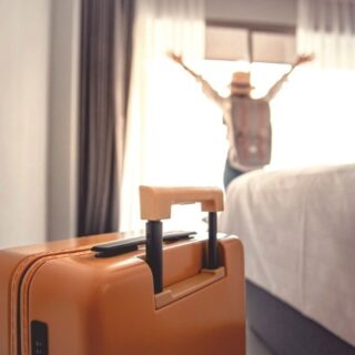 Orange suitcase in the foreground of a hotel room with a person raising their arms by the window in the background, bed on the right side.