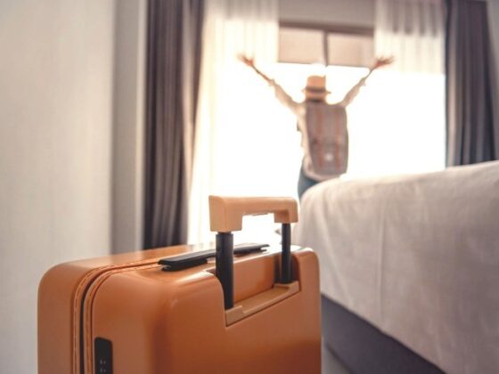 Orange suitcase in the foreground of a hotel room with a person raising their arms by the window in the background, bed on the right side.