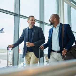 Two businessmen walking through an airport terminal with rolling suitcases, smiling at each other, a plane visible outside the window.