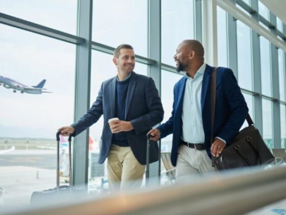 Two businessmen walking through an airport terminal with rolling suitcases, smiling at each other, a plane visible outside the window.