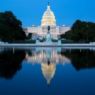U.S. Capitol dome lit at evening, with statue in front and trees, reflected in the calm reflecting pool.