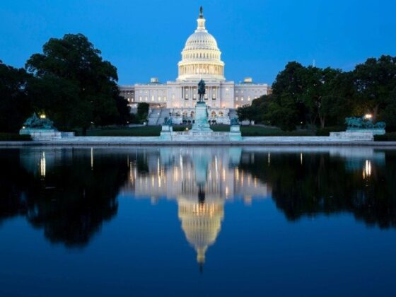 U.S. Capitol dome lit at evening, with statue in front and trees, reflected in the calm reflecting pool.