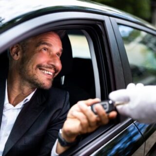 Man in a suit smiles as a car key is handed to him through the driver's window by a gloved attendant.
