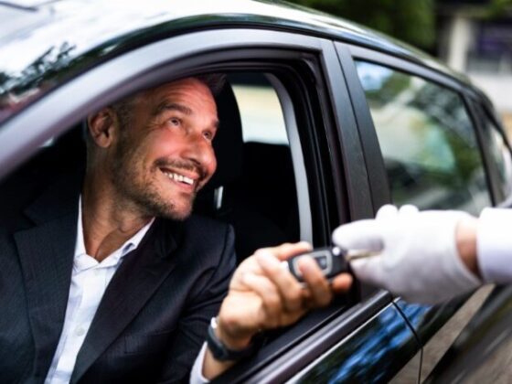 Man in a suit smiles as a car key is handed to him through the driver's window by a gloved attendant.
