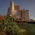 Castle-like beige fortress atop a hill with a landscaped garden in the foreground under a clear blue sky
