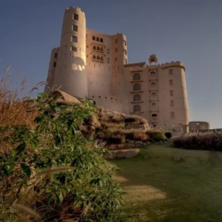 Castle-like beige fortress atop a hill with a landscaped garden in the foreground under a clear blue sky