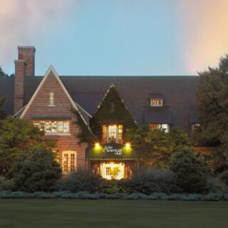 A large brick house with a warmly lit front entrance, surrounded by evergreen shrubs at dusk.