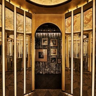 Gold-toned mirrored hallway leading to a small gallery with framed pictures on a dark wall at the end.