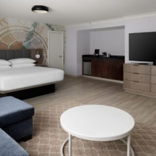 Hotel room with a king bed against a mural wall, a blue sofa, round white coffee table, and a flat-screen TV on a dresser.