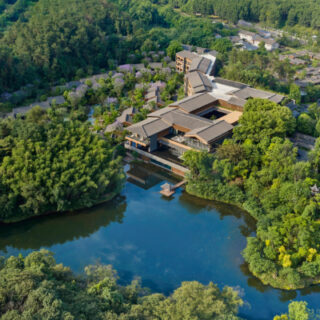 Aerial view of a lakeside eco-resort surrounded by trees, with a large, modern building complex by the water and a dock extending into the lake