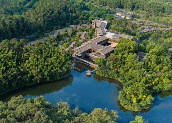 Aerial view of a lakeside eco-resort surrounded by trees, with a large, modern building complex by the water and a dock extending into the lake
