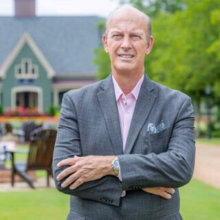 Mature man in a gray suit with arms crossed, posing outdoors in a park with a green house in the background.