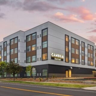 Exterior of a modern four-story Cambria hotel at dusk, with street and trees in foreground.