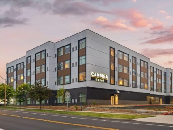 Exterior of a modern four-story Cambria hotel at dusk, with street and trees in foreground.