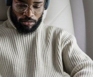Man with a beard wearing a beige ribbed sweater and headphones, seated in an airplane beside a window.