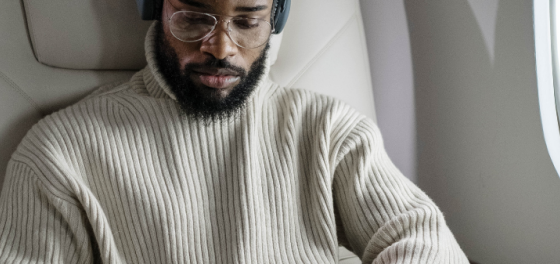 Man with a beard wearing a beige ribbed sweater and headphones, seated in an airplane beside a window.