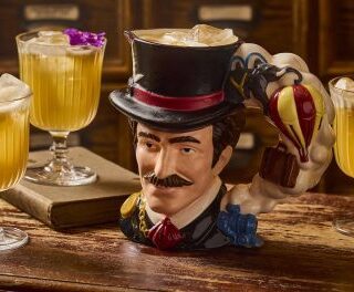 Whimsical top-hat bust mug surrounded by four yellow cocktails on a rustic wooden bar.