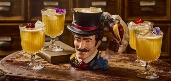 Whimsical top-hat bust mug surrounded by four yellow cocktails on a rustic wooden bar.