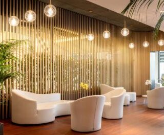 Contemporary hotel lobby with curved white chairs, wooden slat wall, globe pendant lights, and tall potted plants.