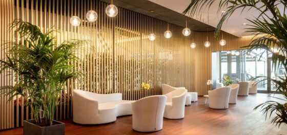 Contemporary hotel lobby with curved white chairs, wooden slat wall, globe pendant lights, and tall potted plants.