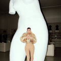 Life-size white sculpture of a torso in a gallery, with a tiny female figure in a fur coat standing on its midsection as if posed.