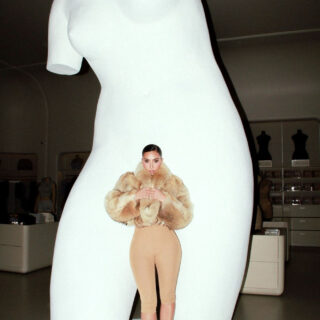 Life-size white sculpture of a torso in a gallery, with a tiny female figure in a fur coat standing on its midsection as if posed.