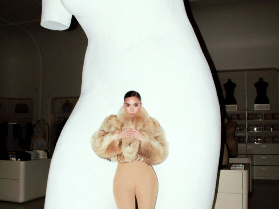 Life-size white sculpture of a torso in a gallery, with a tiny female figure in a fur coat standing on its midsection as if posed.