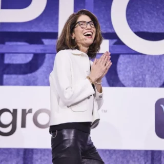 Woman in a white blazer and black pants smiles on stage, hands clasped in front, wearing glasses as she speaks/leads a presentation