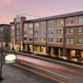 Exterior of a modern brick apartment building along a street at sunset, with lit windows and light trails from passing cars.