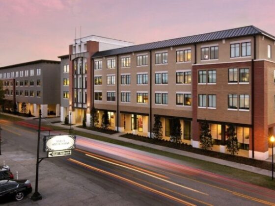 Exterior of a modern brick apartment building along a street at sunset, with lit windows and light trails from passing cars.