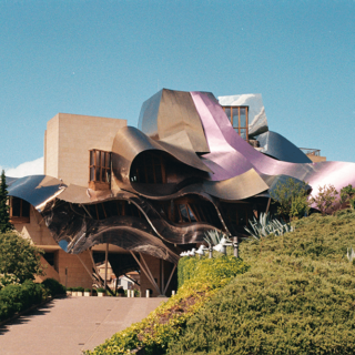 Frank Gehry–designed museum with irregular, flowing titanium panels and a blue sky backdrop, set among green hills.