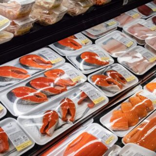 Assorted salmon fillets and other fish cuts arranged in Styrofoam trays in a refrigerated seafood display case with price labels nearby.