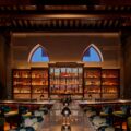 Elegant bar interior with backlit liquor shelves, bottles lined up, and two blue arched windows in the background.