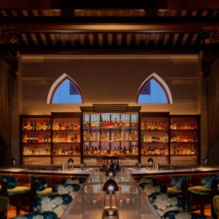 Elegant bar interior with backlit liquor shelves, bottles lined up, and two blue arched windows in the background.