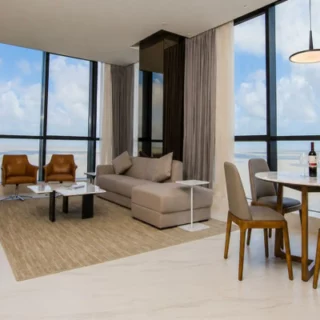 Bright modern living room with large ocean-view windows, beige sofa, tan chairs, and a round dining table with a wine bottle on it.