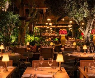 Warm, candlelit restaurant interior with neatly set tables, wine glasses, and lush greenery throughout.