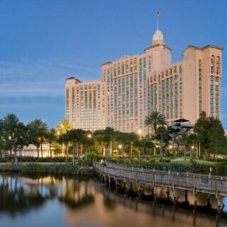 Resort hotel with beige towers, palm trees, and a wooden boardwalk over a calm lake at dusk.