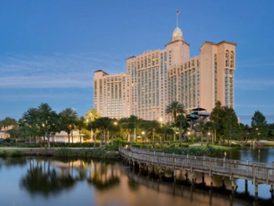 Resort hotel with beige towers, palm trees, and a wooden boardwalk over a calm lake at dusk.