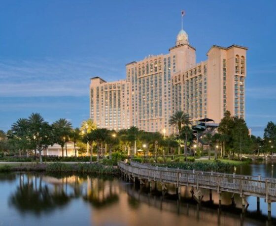 Resort hotel with beige towers, palm trees, and a wooden boardwalk over a calm lake at dusk.