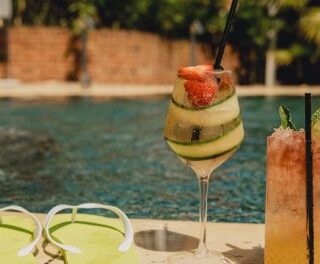 Two colorful cocktails sit on a poolside ledge with bright yellow sunglasses nearby; one tall orange drink with a lime slice and straw, the other greenish with a strawberry garnish.