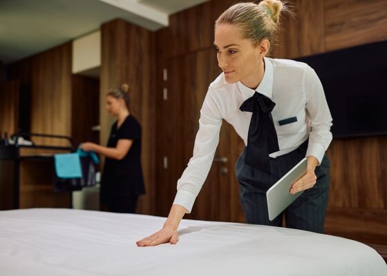 Housekeeper in white shirt and black bow smoothing a bedspread, holding a tablet, in a hotel room with another staff member and cleaning cart in the background