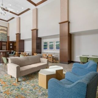 Spacious hotel lobby lounge with a beige sofa, blue armchairs, wooden coffee tables, and a colorful rug underfoot.