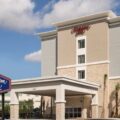 Exterior of a Hampton Inn by Hilton hotel with a covered drop-off entrance, beige and stone facade, and a blue Hampton Inn sign nearby.